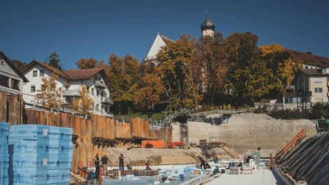Construction site of residential building with workers, insulation materials, and scaffolding during daytime, with trees and a church steeple in the background, showcasing modern building staging and development.