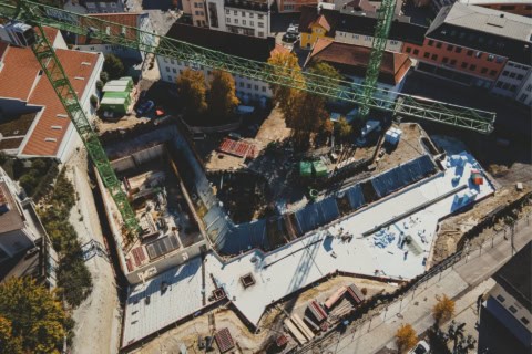 Aerial view of a construction site with a large crane, ongoing building work, and surrounding urban neighborhood with colorful houses and trees.
