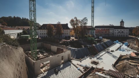 Crane at a construction site in a small town during the daytime, showing ongoing building development with concrete and steel structures, surrounded by trees and colorful buildings.