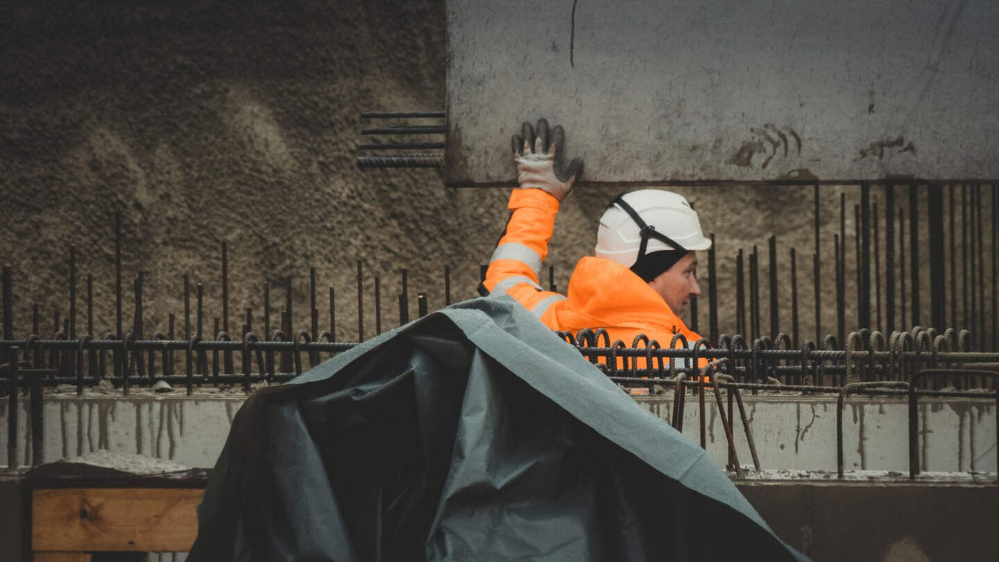 Worker inspecting rebar and concrete formwork at construction site, focusing on structural framing and framing installation, safety gear included.