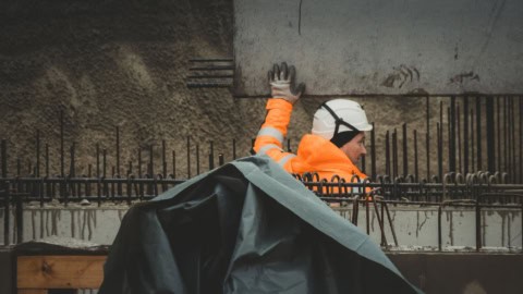 Worker inspecting rebar and concrete formwork at construction site, focusing on structural framing and framing installation, safety gear included.